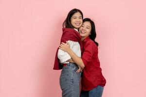 A short brunette Asian woman in a white shirt and a cheerful long-haired girl in a red shirt hug each other on a pink background. photo