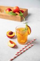 Refreshing peach tea with ice and rosemary. Homemade cold healthy vegetarian drink on a light  background with fresh fruits and paper tube photo