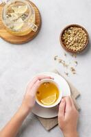 manos participación un vaso taza con manzanilla herbario té en un ligero antecedentes con seco flores y tetera. el concepto de un sano bebida para salud y inmunidad. foto