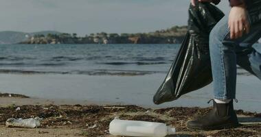 ecoactivist girl collects plastic trash on the coast. Young woman collects plastic garbage in a garbage bag on the sandy beach of the sea. Girl cleans ocean coastline from plastic bottle trash on video