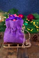 Canvas purple bag with red gift box on sled on wooden boards. Spruce branches, toys and defocused lights. Christmas, New Year, sale. Soft focus. Evening. Copy space photo