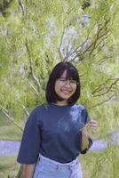 asian teenager toothy smiling and standing under green leaves tree photo