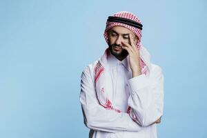 Pensive muslim man in thobe and ghutra headscarf standing in thoughts and holding temple. Arab person wearing traditional clothes looking away posing in studio with thoughtful face expression photo