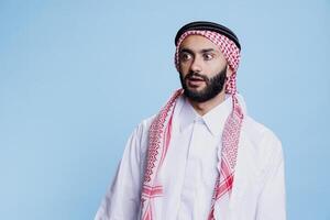 Man wearing traditional muslim clothes looking away shocked face expression. Arab person dressed in thobe and headscarf standing with surprised emotion and open mouth while posing in studio photo