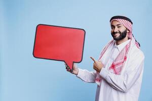 Smiling muslim man holding red speech bubble mockup and pointing with finger portrait. Person dressed in arab traditional clothes standing with message banner and looking at camera photo