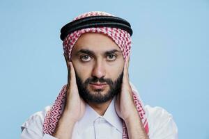 Muslim man closing ears with palms while showing hear no evil three wise monkeys concept studio portrait. Arab person wearing traditional clothes showcasing hearing problems and looking at camera photo