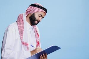 Man wearing traditional islamic thobe and headscarf clothes writing in clipboard, filling application form. Arab dressed in checkered ghutra and robe taking notes in checklist while posing in studio photo