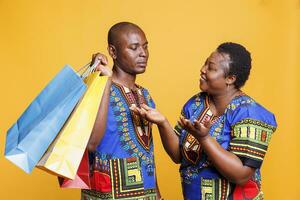 Smiling african american woman enjoying shopping spree while man carrying heavy paper bags. Cheerful wife happy with purchase while bored husband holding paperbags on shoulder photo