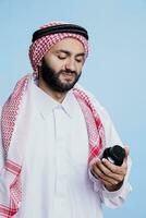 Man wearing traditional muslim attire checking instruction on supplement bottle with unhappy expression. Arab dressed in thobe and ghutra holding medication for disease treatment photo