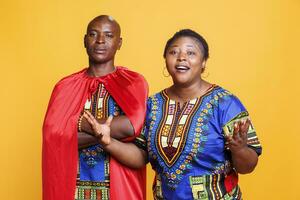 Confident african american man wearing superman cape standing with arms crossed while excited woman looking at camera. Serious guy dressed in superhero cloak posing with cheerful wife photo