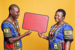 Smiling couple holding red speech bubble mockup and showing with hand. Cheerful african american man and woman standing with blank banner with copy space for promotion message photo