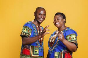 Smiling african american man and woman wearing ethnic clothes showing thumb up and okay gesture portrait. Couple giving positive feedback and looking at camera on studio background photo