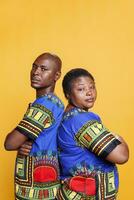Serious black couple dressed in ethnic clothes posing with arms crossed studio portrait. Confident man and woman standing back to back with folded hands and looking at camera photo