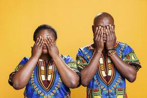 African american couple covering eyes with arms, showing hear no evil three wise monkeys portrait. Black man and woman pair holding palms on face on orange studio background photo