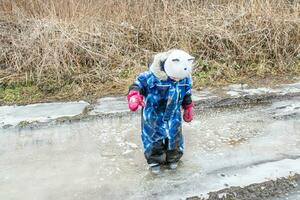 A small child in a blue jumpsuit stands in rubber boots in a puddle in the spring after the snow melts photo