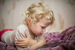 un pequeño niña es acostado en un sofá con un tableta. el niño relojes dibujos animados en lugar de juegos y desarrollo en artilugio. un solitario niño en línea foto