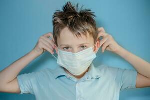 boy puts on a medical protective mask on blue background. child with flu, influenza or cold protected from viruses, pollution in bad epidemic situation, among patients with coronavirus photo