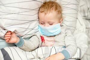 pequeño niño duerme en cama en protector médico mascarilla. enfermo niño en blanco cama. niño niñito uno años antiguo con gripe, influenza o frío protegido desde virus, contaminación entre pacientes con coronavirus foto