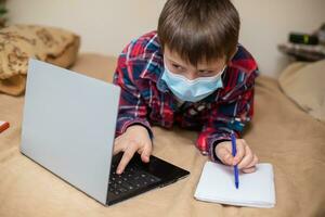 boy in protective medical mask lies near laptop and writes with left hand in notebook. child does lessons watching online broadcast lessons on Internet. e-learning, remote studying internet concept photo