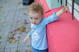 alegre pequeño hermosa niña soportes cerca un rojo pared. el niño elevado su mano caminando abajo el calle. foto