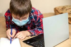 boy in protective medical mask lies near laptop and writes with left hand in notebook. child does lessons watching online broadcast lessons on Internet. e-learning, remote studying internet concept. photo