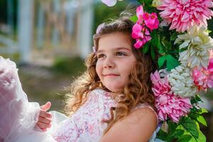 hermosa pequeño niña de preescolar años en un delicado vestir sentado en un columpio decorado con flores . foto