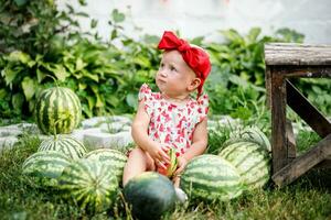 Portrait of a girl looking up in surprise in a red headband sitting near watermelons at the fair. Fresh ripe green watermelons on the grass on a sunny summer day photo