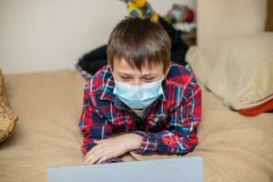 teenage boy in protective medical mask coughs in fist. child remotely does lessons lying on bed near laptop. Online training during coronavirus quarantine. Home education photo