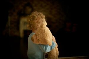 Little girl with curly hair holds in hands of teddy bear. The child is on the dark room with his toy. Soft focus photo