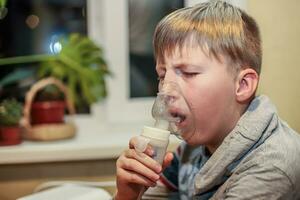 A boy is being treated by inhaling medicine through a nebulizer at home in the kitchen by the window photo