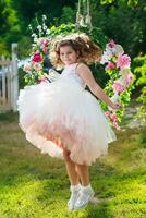 Beautiful little girl of preschool age in delicate dress Jumps near swing decorated with flowers . photo
