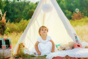 A girl in a white dress sits in a white tent on a background of green bushes. The child sat on his haunches. photo