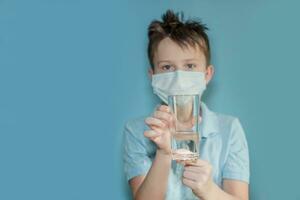 A boy in a protective medical mask holds a glass of water. Compliance with the drinking regime. Fighting coronavirus. child with flu, influenza or cold protected from viruses photo