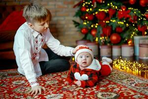 hermano con pequeño hermana a el nuevo año árbol. familia celebra Navidad. 6 6 meses antiguo pequeño chico aprende a gatear foto