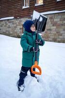 A boy in a green down jacket with a shovel stands in the yard in winter. photo