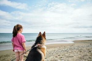 un niña en un rosado camiseta y un perro Mira a el mar. foto
