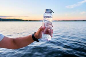 A man holds a bottle of water against the backdrop of a lake. photo