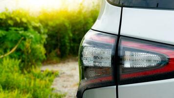 Rear side view of the silver taillights of a parked silver sedan. It is parked in an open area surrounded by green trees. The weather is bright and sunny during the daytime. photo