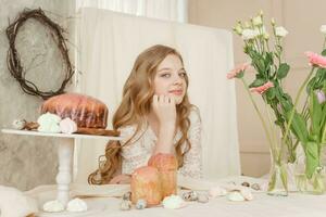 un niña con largo pelo en un ligero vestir es sentado a el Pascua de Resurrección mesa con pasteles, primavera flores y codorniz huevos. contento Pascua de Resurrección celebracion. foto