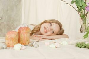 un niña con largo pelo en un ligero vestir es sentado a el Pascua de Resurrección mesa con pasteles, primavera flores y codorniz huevos. contento Pascua de Resurrección celebracion. foto