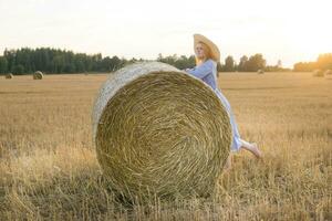 A red-haired woman in a hat and a blue dress walks in a field with haystacks. photo