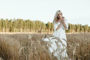 A blonde woman in a long white dress walks in a wheat field. The concept of a wedding and walking in nature. photo