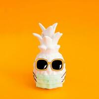 Pineapple in a medical mask and sunglasses on orange background. Summer in Covid-19. Vision for the summer of 2020 during the coronavirus pandemic. photo