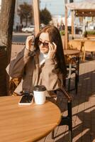 un elegante morena mujer en Gafas de sol es sentado a un mesa en un al aire libre café con café y un teléfono. el niña es hablando en el teléfono. otoño caminar foto