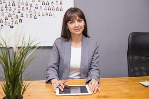 A brunette businesswoman in a gray jacket at her desk with a tablet in her hands. Business portrait in the office. photo