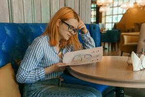 A woman in a cafe at a table with a laptop is studying the design of a person. The concept of studying esoteric sciences. A red-haired woman with glasses studies a rave card by human design. photo