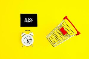 A white alarm clock, a shopping cart, and a black square that says black Friday on a yellow background. The topic of sales. photo