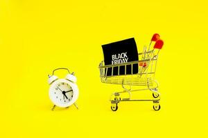 A white alarm clock, a shopping cart, and a black square that says black Friday on a yellow background. The topic of sales. photo