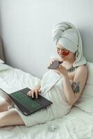 Tver, Russia-august 2, 2021 A woman after a shower is lying on the couch with a laptop and an eye mask at the same time. The concept of multitasking in the twenty-first century. photo