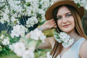 Beautiful young girl in white dress and hat in blooming Apple orchard. Blooming Apple trees with white flowers. photo